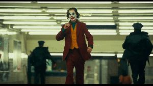 Joker - Arthur has his face painted white, with blue diamonds over his eyes and red big lips. His hair is dyed green and hangs down his face. He is dressed with a red suit, yellow vest, green shirt. He smiles as he brings his hand to his mouth. Police run in the opposite direction behind him
