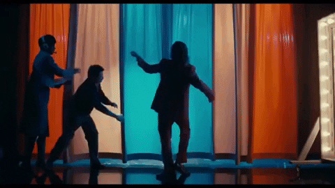 Joker is dancing in a dark suit as bright curtains to the TV show are lifted up