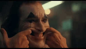 Arthur has his face painted white with blue triangles above and below his eyes. He uses his fingers to force his mouth into a grotesque smile