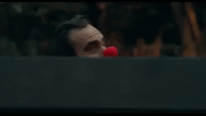 Joker is dressed in white face make up with a large plastic red nose. He is swaying side to side and walking behind a large fence outside the Wayne estate