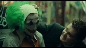 A White man with curly hair pulls Arthur's wig off his head. Arthur has his face painted like a clown. He looks upset and his arms are close to his side
