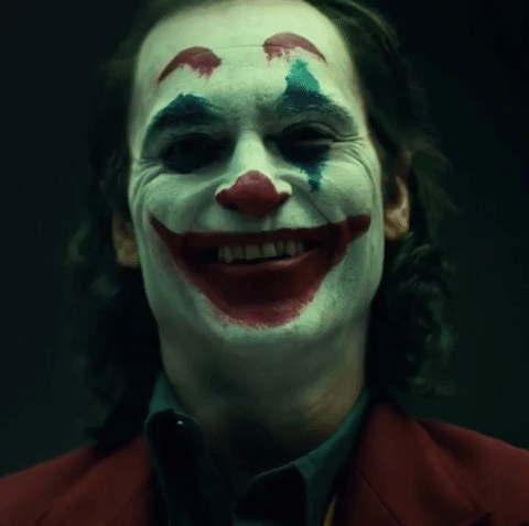 Arthur is in full Joker make-up. His face is painted white with crass red lips overdrawn over half his face. He has large red eyebrows painted high on his forehead. His eyes are framed by two blue triangles. He smiles and raises his eyebrown as he exhales