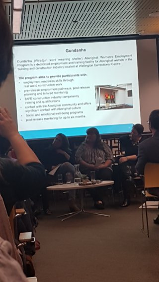 Panellists sit at the front of a large room. The screen projects a PowerPoint slide with a photo of women in high-visibility vests dressed for construction. The slide describes the program and its aims to provide training, accreditation and support for women prisoners