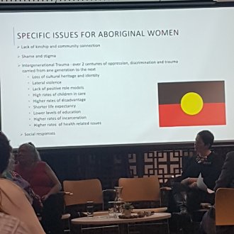 Panellists sit at the front of a room with projected PowerPoint slide behind them. The text say: lack of kinship and commmunity connection; shame and stigma; Intergenerational trauma - over 2 centuries of oppression, discrimination and trauma carried from one generation to the next; and social responsesst is described in my blog post