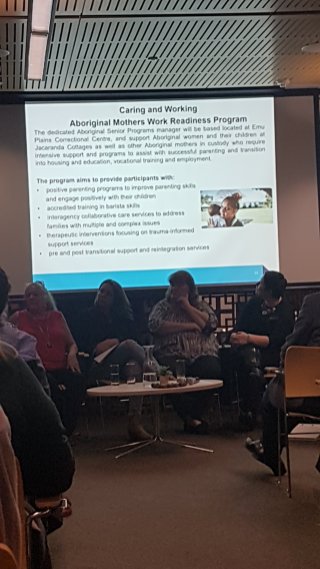 Panellists sit in the front of the room with a PowerPoint slide projected behind them. The slide shows an Aboriginal woman holding a baby. The text is hard to read without zooming, but it describes the program and aims