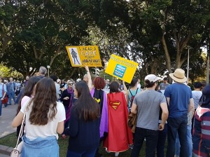 Protesters gathered in Hyde Park, Sydney. Two women are dressed in capes. They are holding yellow signs. One says