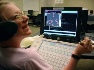 A White disabled woman sitting in her wheelchair. She has her face turned backward and to the side, smiling at someone outside the shot. Her communication device is in front of her, which includes a large keyboard