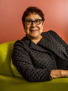 Prof Aileen Moreton-Robinson is smiling. She wears black glasses and sits on a lime-green couh