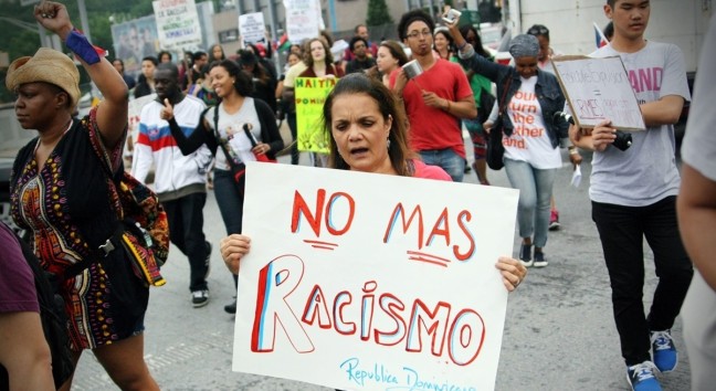 Protesters hold signs on the str4eet. One Brown woman is chanting and her sign says: No mas racismo Republica Dominicana