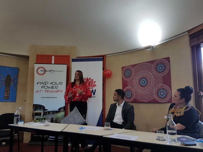 Stewart is standing and talking animated, her co-panellists look up at her as she speaks. Various banners and Aboriginal artworks hang in the background