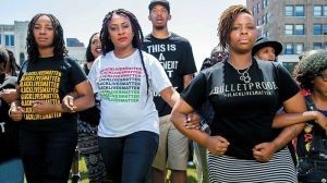 The three #BlackLivesMatter co-founders are walking with their arms interlocked. They stare with determination at the viewer. Tometi on the left wears a black BLM t-shirt. Garza in the cetre wears a white BLM t-shirt. Cullors on the right wears a t-shirt that says 'bullet proof.'