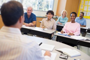 Photo depicting a classroom. A White man sits at the front with his back to the viewer. Five people of various racial backgrounds can be seen smiling, sitting opposite the White man, appearing to be his students