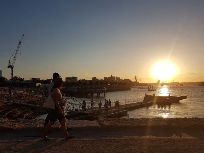 Two people walk along the foreshore. In the background, other groups gather at the pier taking photos and looking around, while others are near a giant vessel. The sun shines brightly as it sets over the water