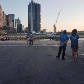 Tree Sydney Festival volunteers stand together to the left hand side, a couple can be seen with their arms around each other walking in the background. The Barangaroo foreshore is seen in the far disance with buildings towering over the top