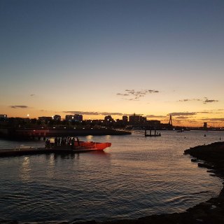 Landscape view of Barangaroo foreshore, with water surrounding the lit vessel in the background, and figures huddled in front of it. In the background are buildings and a construction site crane high above. It is starting to get dark and the sunsets beautifully in the far distance