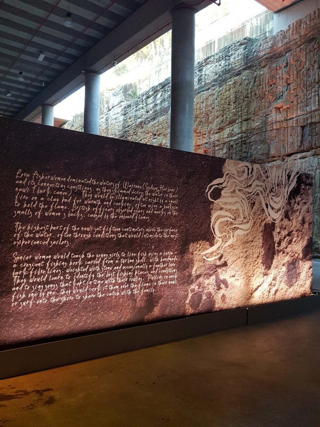 A beautiful mural with photos of Country with stylised drawing of the water on the rick, over rocks. Cursive writing on the right reads: Eora fisherwomen dominated the waters of Warrane (Sydney Harbour) and its connecting coastlines, as they skimmed across the water in their nawi (bark canoe). They would be illuminated at night by a small fire on a clay pad for warmth and cooking, often using a banksia to hold the flame. British officers observed scars and marks on the smalls of women's backs, caused by the onboard flames. The highest part of the nawi sat fifteen centimeters above the surface of the water, often through conditions that would intimidate the most experienced sailors. Senior women would teach the young girls to line fish using a burra - a crescent fishing hook carved from a turban shell, with handmade bark fibre lines, weighted with stone and occassionally a fether lure. They would learn to identify the best fishing places and conditions and to sing songs that kept time with their rowing. Pulling in each fish one by one, they would cook it over the flame in their nawli or surf onto the shore to share the catch with the family