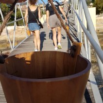 Close-up of my wooden bucket. Part of my hand is seen at the top of the photo, and other people walk across a bridge in the background. It is a sunny day