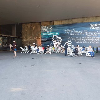 The exterior of Barangaroo undercover area. In the background is a giant mural with the Sydney Festival logo and the title "Four Thousand Fish." Information about the exhibition can be seen in cursive writing but cannot be easily read from a distance. People sit on white deck chairs, while others walk in either direction