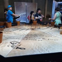 A light-coloured hexagon-shaped wooden table made of interlocking triangles and black checkered lines. Wooden buckets sit on the corners. People are smiling in hte background, carrying their ice fish moulds into the portable freezers