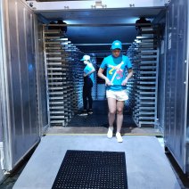 A young person looks down as they walk out of a giant portable freezer. In the background a woman stands in the middle of metal racks, awaiting the ice fish