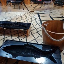 Part of a round table can be seen with black drawing imitating fishing lines. On top sits my wooden bucket and fish mould. In the background, children and adults look down at their own frozen fish