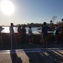 People stand in front of the Four Thousand Fish installation by the shore. A giant metal vessel (coolamon) holds hundreds of ice fish. The sun is shining high on the left hand side. Some people stand close and lean in to take photos