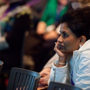A woman of colour is sitting, her body is forward and attentive. She rests her chin on her hand and appears to be concentrating