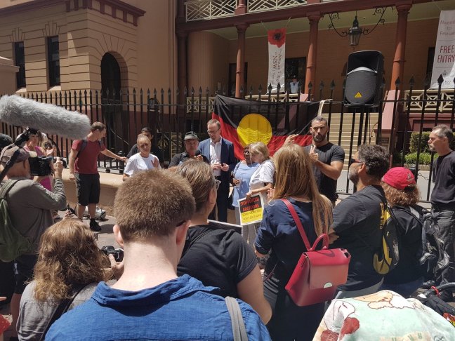 Protesters gather out the front of the Parliament of New South Wales. A lage Aboriginal flag hangs over the gates. An Aboriginal man is signing translation in Auslan. There are camera crews and photographers.