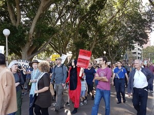 People march across Hyde Park, Sydney. A man dressed in a furry black and red onesie suit holds a red sign that reads: Heed our warning signals