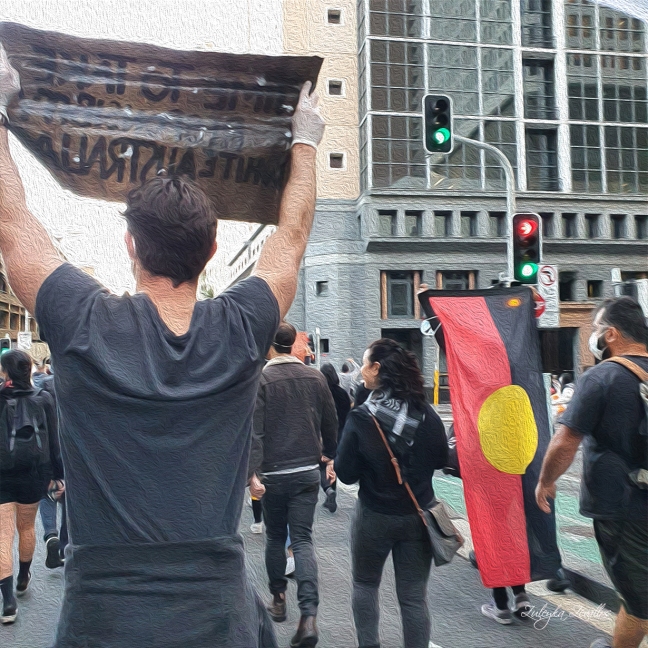 People march during the Black Lives Matter protest in Sydney. One man holds up a sign. Another person holds up a large Aboriginal flag