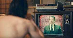 Arthur is shirtless, with his back to the audience. He is watching Thomas Wayne, a White man in a suit, speaking on television