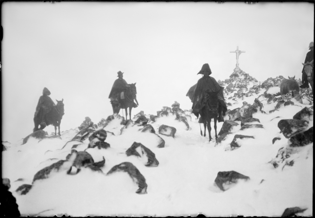 A blurry photo of three Quechuan people in a snowy mountain. A large cross is in the far background