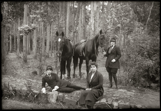Mestiza women are dressed in modern 1920s riding gear. Two are sitting and laying on the ground. Another woman stands next to two horses