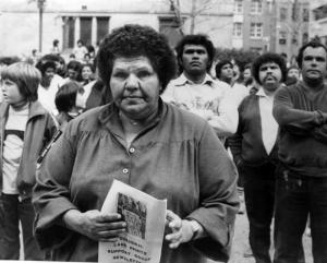Black and white photo of Coleen Shirley Perry Smith stares with determination at the audience. She is dressed in a large shirt. She holds a poster that reads: Aboriginal land rights support group newsletter. Other Aboriginal people stand in the background