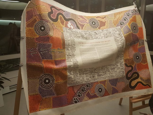 Close-up of the Uluru Statement. It is a beautiful, large cloth document that has been signed by prominent Aboriginal leaders from around the country. The large border is colourful Aboriginal art of various orange and brwn colours. The Statement is reproduced in full in the centre, surrounded by signatures