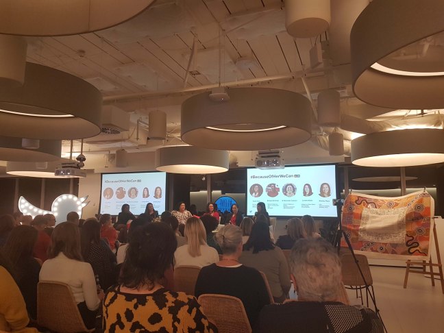 Photograph from the back of the room captures a lage seated crowd. The panellists sit in the front with two large screens showing their Twitter handles