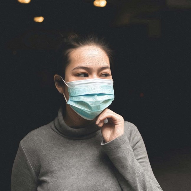 An Asian woman wears a surgical mask. She's touching her hand to the bottom of her chin as she looks off to the side
