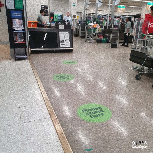 Entrance to a supermarket. Stickers on the ground say "please stand here." Workers are busy in the background
