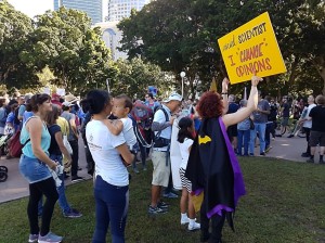 People are gathered at Hyde Park in Sydney. A woman of colour holds a young child in her arms. In front of her a White woman dressed in a superhero cape holds a yellow sign that reads: