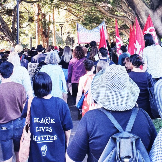 Protesters march through Sydney. One of them wears a tshirt that says "Bla(c)k lives matter." Many carry communist flags. A large banner in front has the colours of the Aboriginal flag