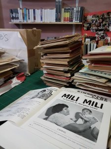 A stack of books and periodicals are shown on a table, with the black and white magazine, Mili Mili, in the foreground. The cover has an Aboriginal woman holding a baby up in the air