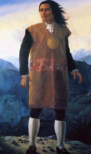 Painting of Tupac Amaru. He is a Quechuan man with Brown skin. He wears a large gold medallion of the sun god Inti, and a traditional dress with modern shoes