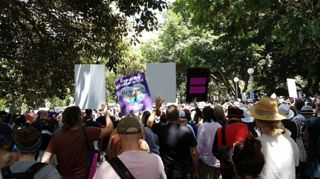 Amongst a packed crowd of able-bodied people standing, a sign reads "Demand a better world for women and girls"